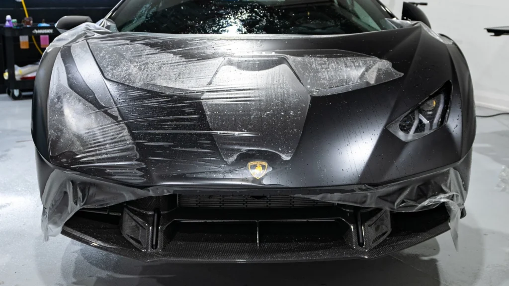 A black Lamborghini undergoing matte paint protection film (PPF) installation in a modern studio, with professionals applying the film under bright, focused lighting.